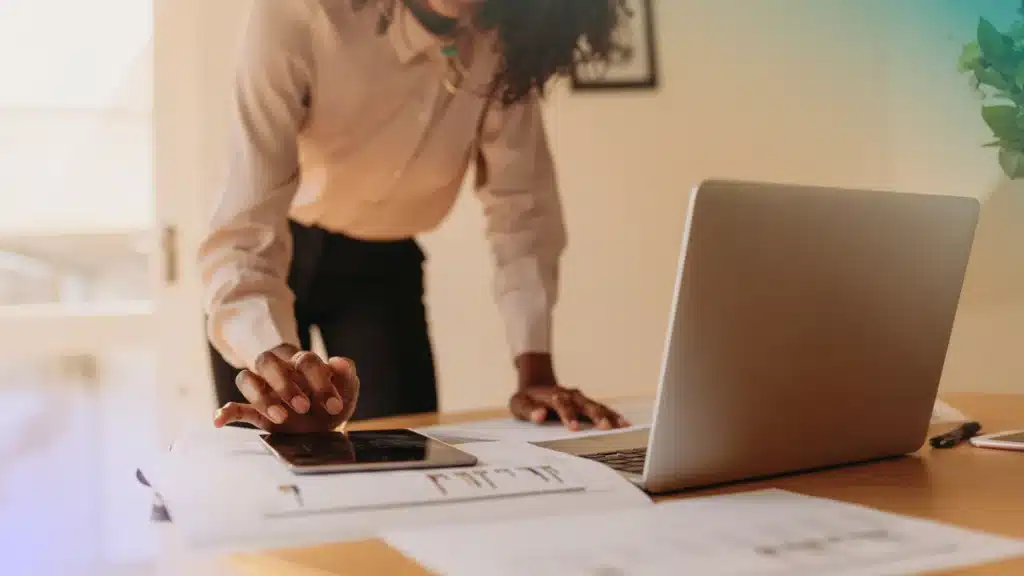 Black woman dressed in business attire at a desk standing, typing on a tablet, and in front of a computer. She is smiling, but you cant see all of her face