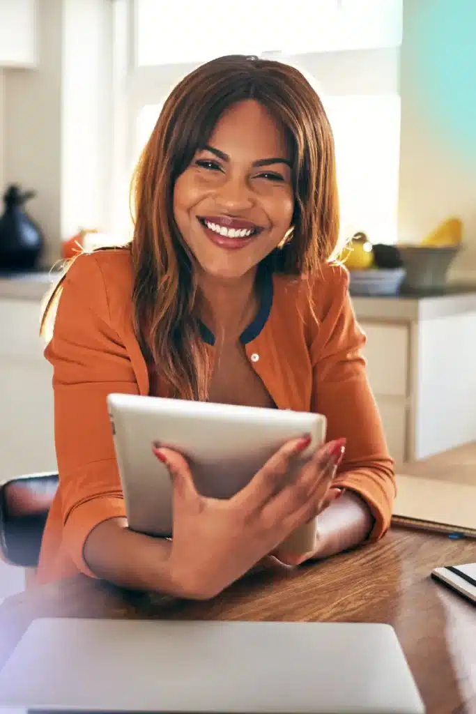 attractive black female with a tablet in her hands. She is smiling at the camera, and appears to be at a kitchen table working